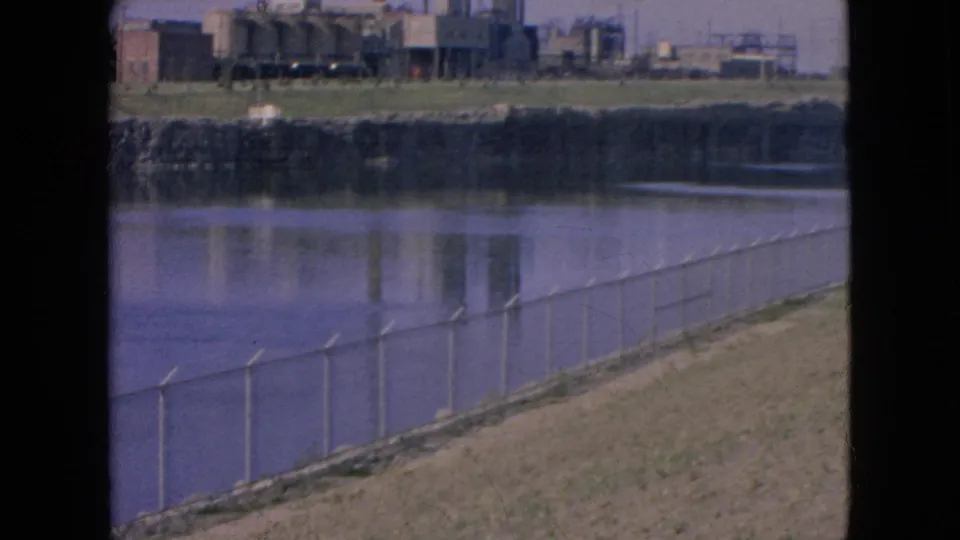 1964 Niagara Falls, New York frame showing a diagonal fence along the riverbank with industrial buildings and reflections across the water under clear light.