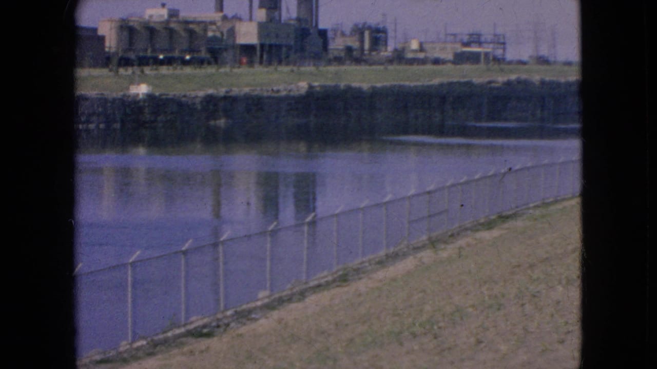 1964 8mm view in Niagara Falls, New York, showing a chain-link fence along a sloped riverbank with industrial buildings and smokestacks across the water.