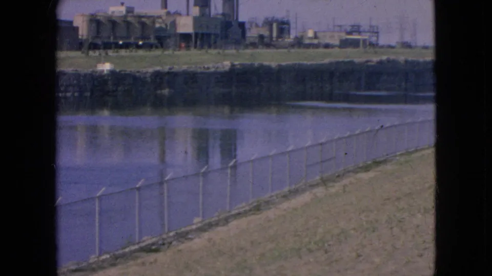 1964 8mm view in Niagara Falls, New York, showing a chain-link fence along a sloped riverbank with industrial buildings and smokestacks across the water.