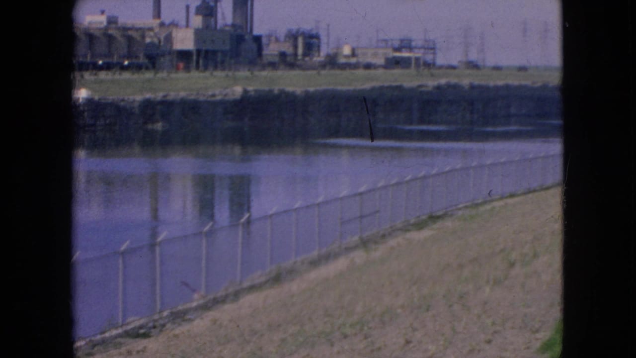Factory complex and river viewed past a fence and dark vertical frame.