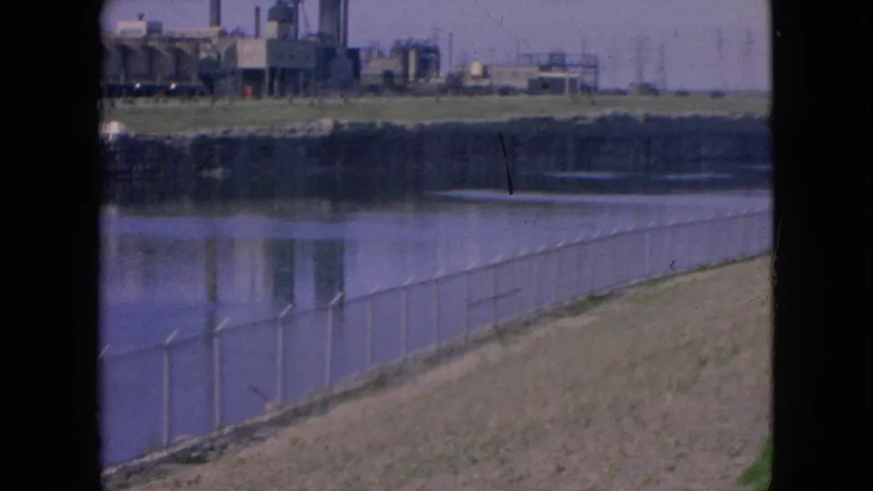 1964 Niagara Falls 8mm frame with a dark vertical border, calm river water, chain-link fence, and distant factory structures under a hazy sky.