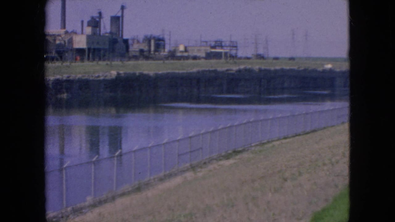 1964 home-movie frame from Niagara Falls showing a fenced riverbank, reflective water, distant industrial buildings, and power infrastructure softened by 8mm grain.