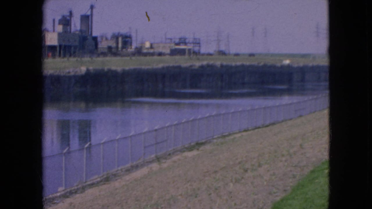 1964 8mm view of a Niagara Falls river edge with chain-link fencing, a grassy bank, calm water, and distant factory buildings beneath a pale sky.