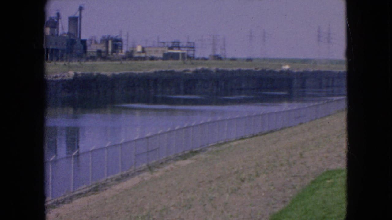 1964 8mm view in Niagara Falls, New York, showing a fenced riverbank, calm water, grass, and distant industrial buildings under a hazy sky.