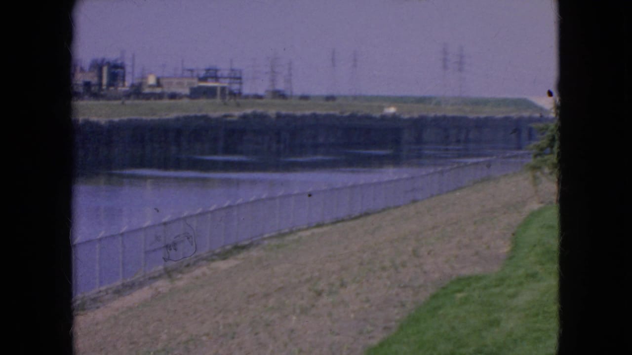 1964 Niagara Falls home-movie frame with a chain-link fence along a river, grassy bank in the foreground, and industrial buildings across the water.