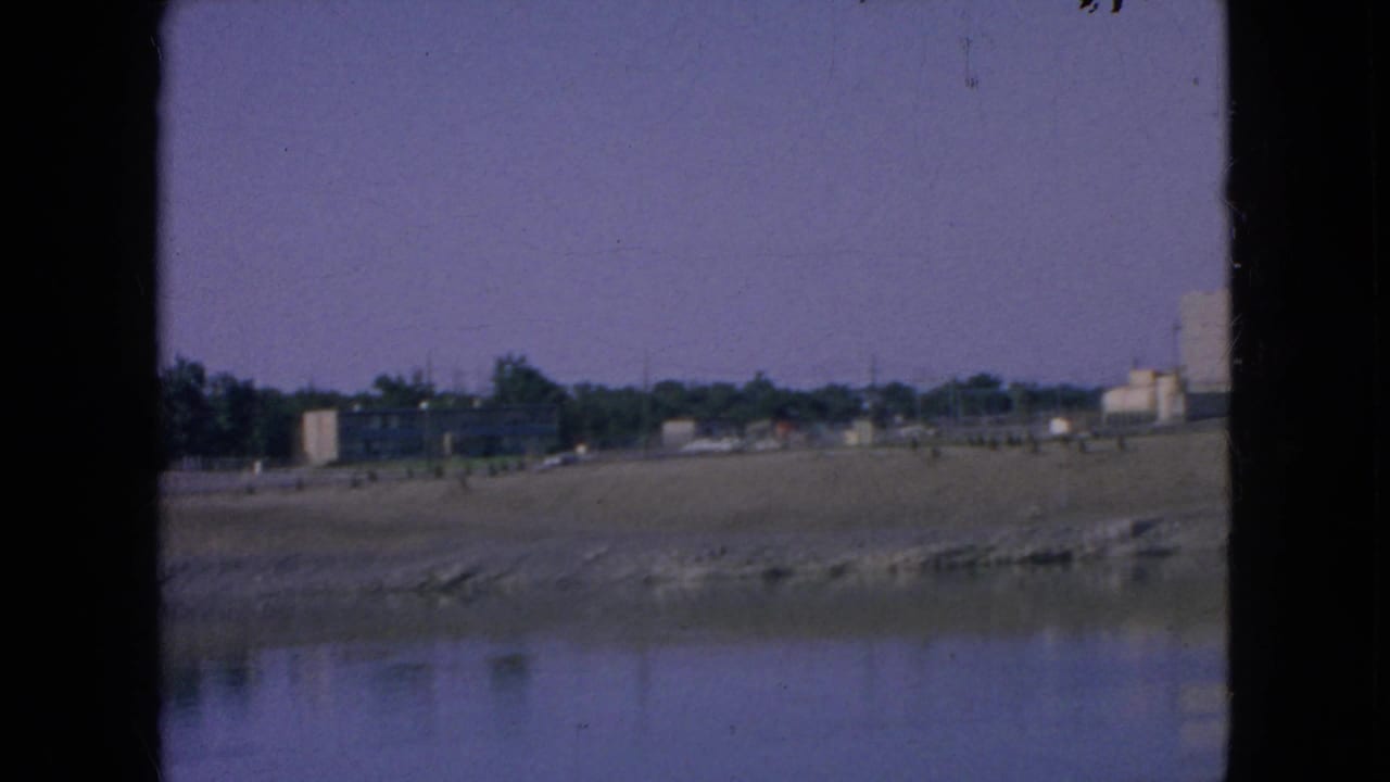Wide 1964 8mm view across water toward a low shoreline, tree line, small structures, and industrial buildings at the right edge.