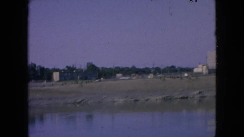 Wide 1964 8mm view across water toward a low shoreline, tree line, small structures, and industrial buildings at the right edge.
