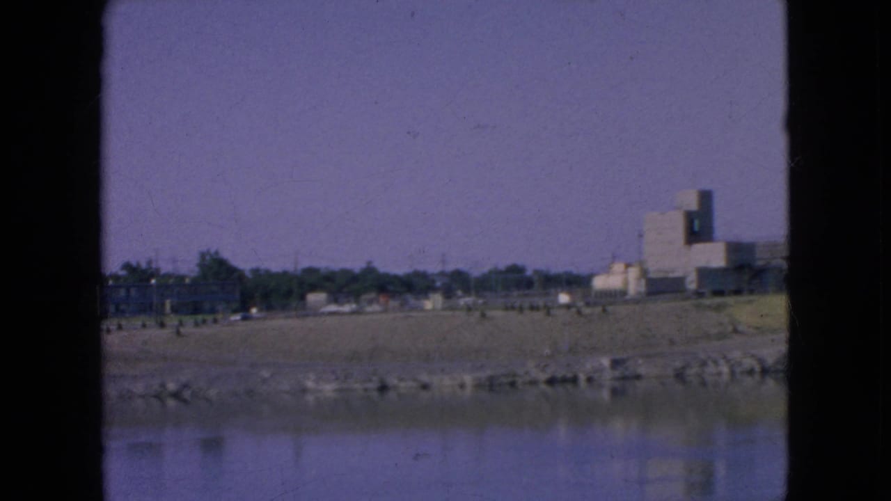 1964 Niagara Falls frame looking across a calm waterway toward a sandy or gravel riverbank, distant trees, and industrial buildings on the right.