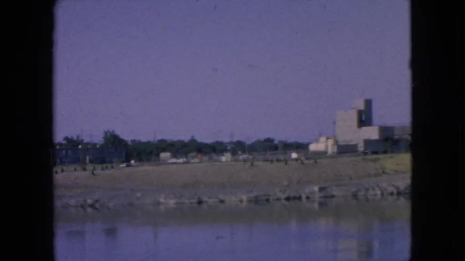 1964 Niagara Falls frame looking across a calm waterway toward a sandy or gravel riverbank, distant trees, and industrial buildings on the right.