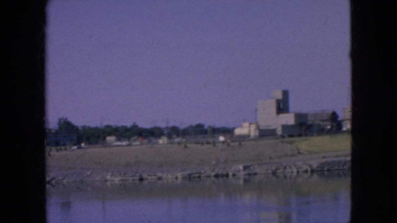1964 8mm frame near Niagara Falls, New York, showing calm water, a stone riverbank, grassy open ground, and distant modern rectangular buildings under a purple-tinted sky.
