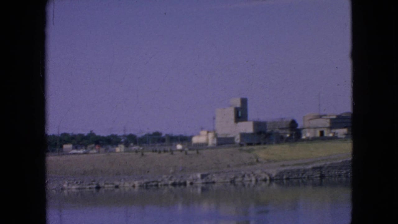 1964 8mm frame near Niagara Falls, New York, with calm river water in the foreground, a sloped stone embankment, open grass, and several distant buildings on the far bank.