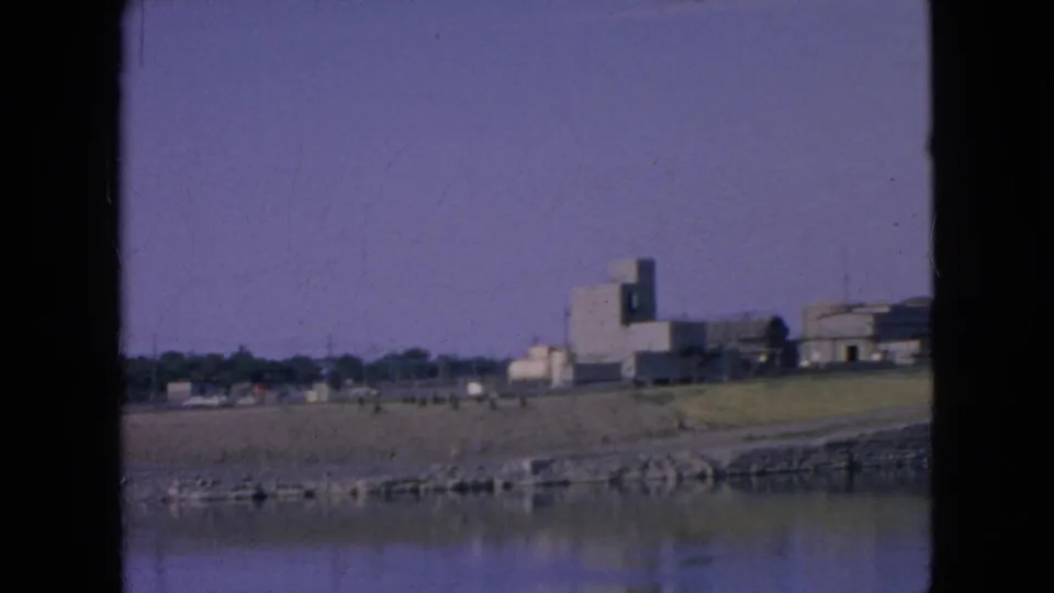 1964 8mm frame near Niagara Falls, New York, with calm river water in the foreground, a sloped stone embankment, open grass, and several distant buildings on the far bank.