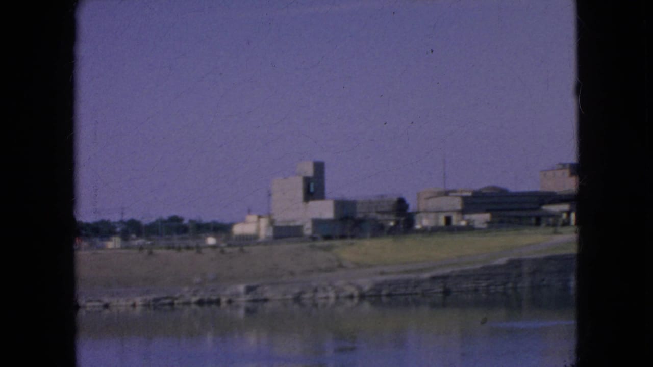 1964 8mm frame near Niagara Falls, New York, showing a quiet riverside field, stone embankment, reflective water, and a cluster of low modern buildings in the distance.
