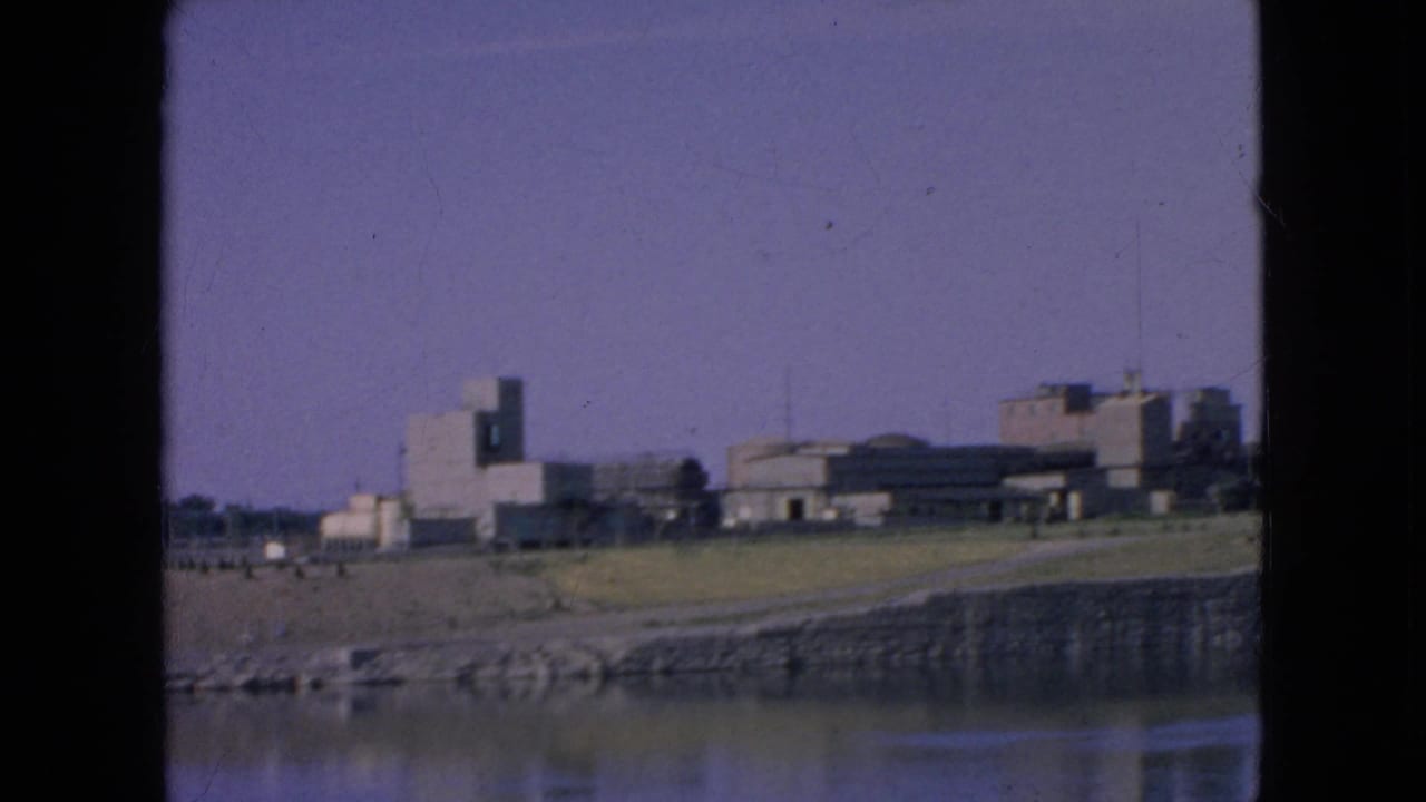 1964 8mm frame near Niagara Falls, New York, capturing calm water, a stone-edged bank, grassy ground, a curving road or path, and distant modern buildings under a clear sky.