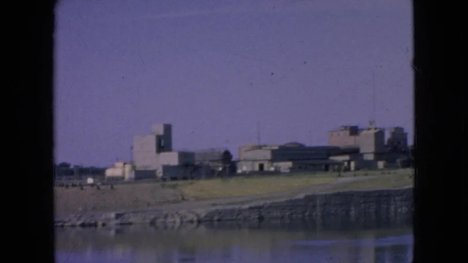 1964 8mm frame near Niagara Falls, New York, capturing calm water, a stone-edged bank, grassy ground, a curving road or path, and distant modern buildings under a clear sky.