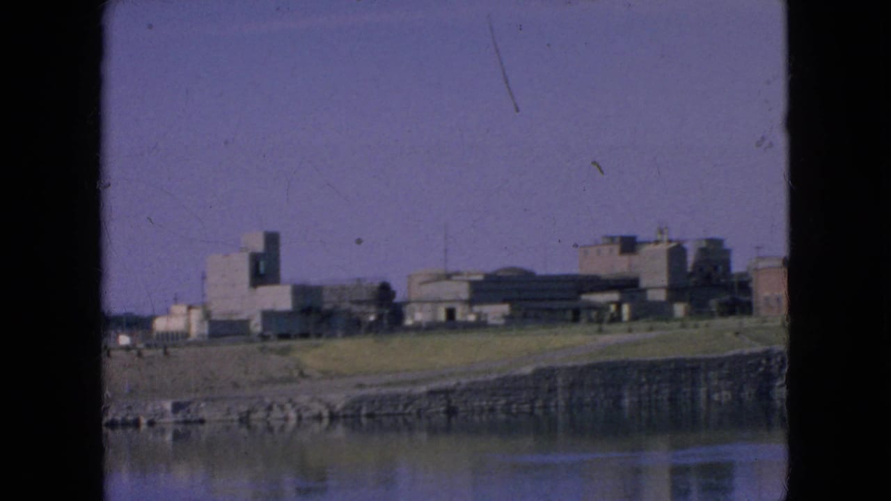 1964 8mm frame showing a distant exterior view of Alcatraz across San Francisco Bay, with low buildings, shoreline, water reflection, and dark gate edges.