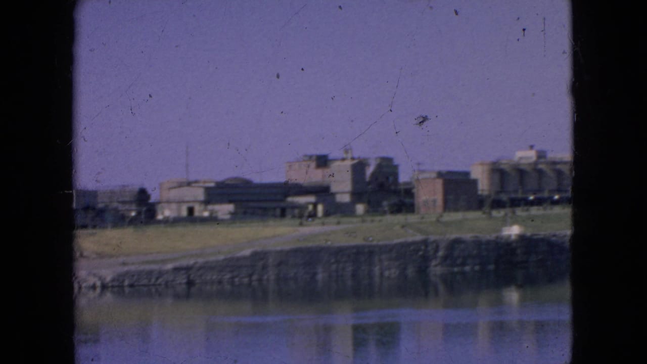 1964 home-movie frame with a distant Alcatraz exterior, shoreline wall, bay water reflection, and soft 8mm color shift.