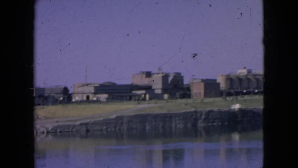 1964 home-movie frame with a distant Alcatraz exterior, shoreline wall, bay water reflection, and soft 8mm color shift.