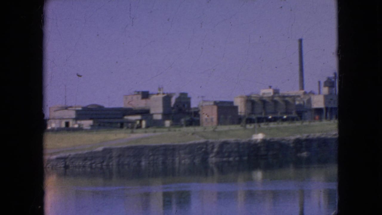 Distant 1964 8mm frame of Alcatraz across San Francisco Bay, showing the prison complex, shoreline, water reflection, and a tall smokestack at the right.