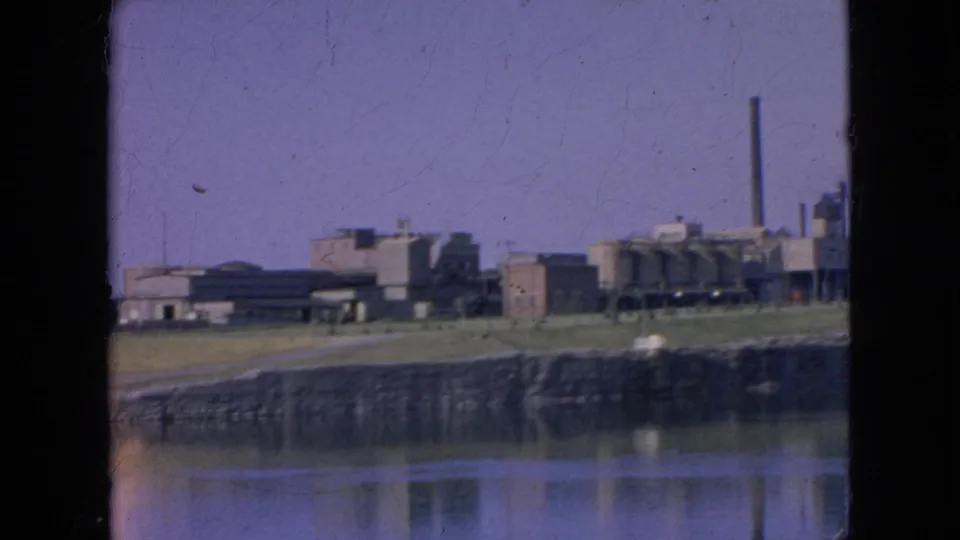 Distant 1964 8mm frame of Alcatraz across San Francisco Bay, showing the prison complex, shoreline, water reflection, and a tall smokestack at the right.