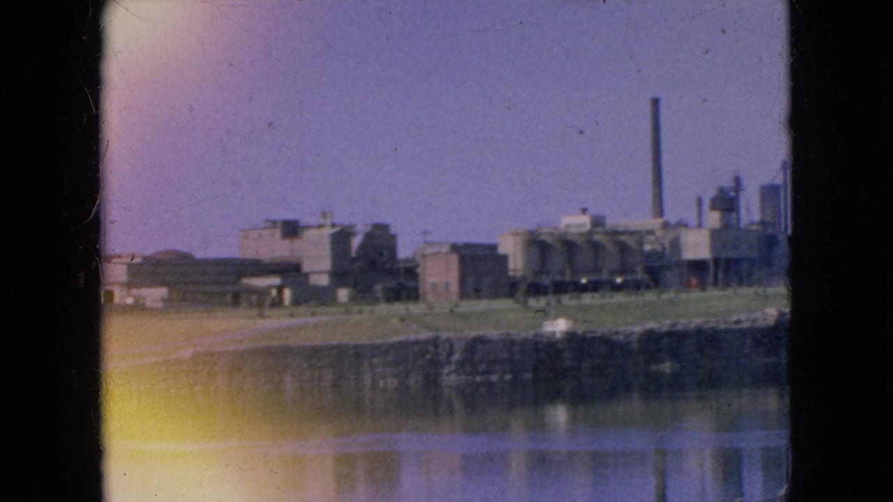 1964 8mm frame showing a distant industrial plant near water in Niagara Falls, New York, with a tall smokestack and reflected shoreline.