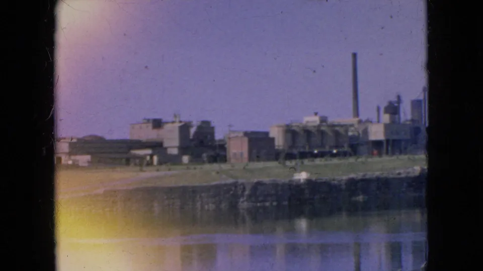 1964 8mm frame showing a distant industrial plant near water in Niagara Falls, New York, with a tall smokestack and reflected shoreline.