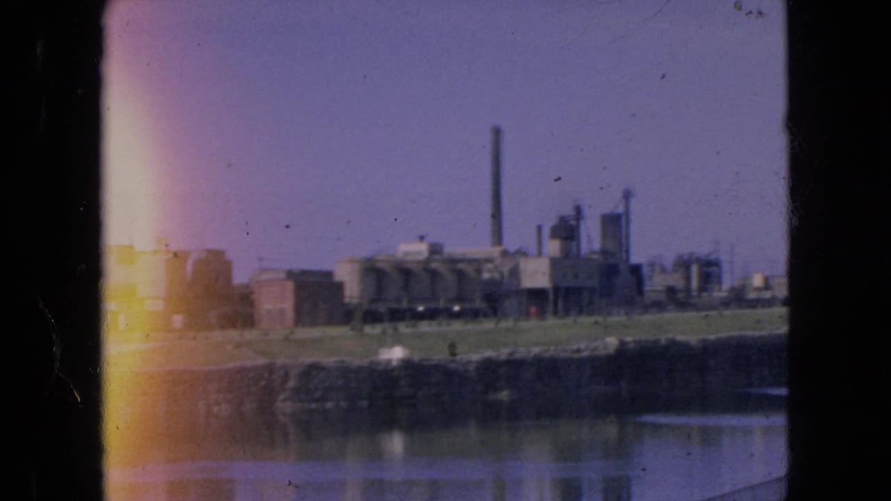 1964 home-movie frame of an industrial plant beside water, with a tall stack, low buildings, shoreline, reflections, and left-edge light flare.