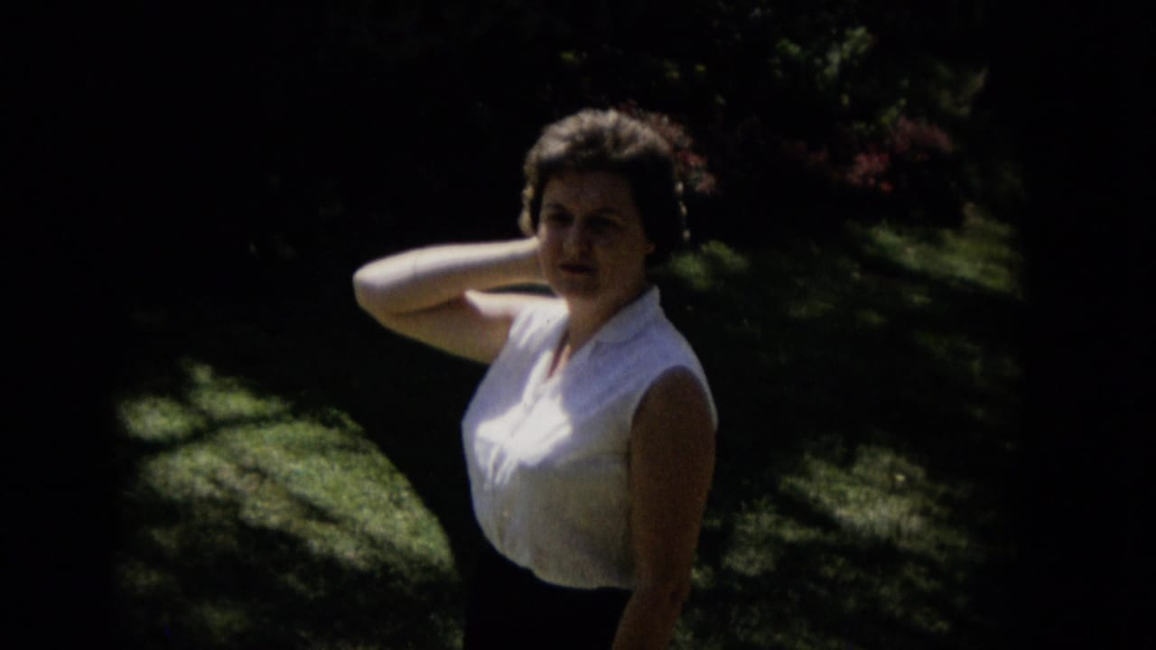 In 1965 Hawaii home-movie footage, a woman in a white sleeveless blouse and dark skirt poses on a sun-dappled lawn with heavy garden shade around her.