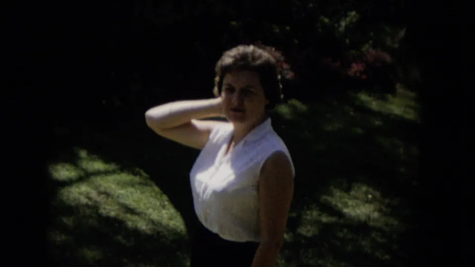 In 1965 Hawaii home-movie footage, a woman in a white sleeveless blouse and dark skirt poses on a sun-dappled lawn with heavy garden shade around her.