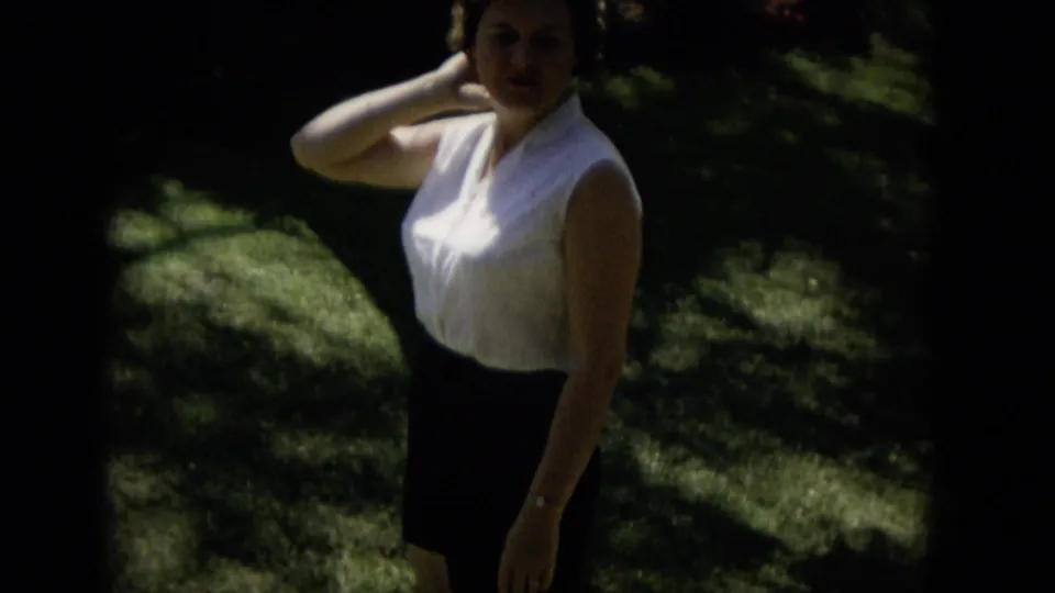 The woman remains posed outdoors in the 1965 Hawaii home-movie scene, partly obscured by shadow and soft focus as sunlight falls across the grass behind her.