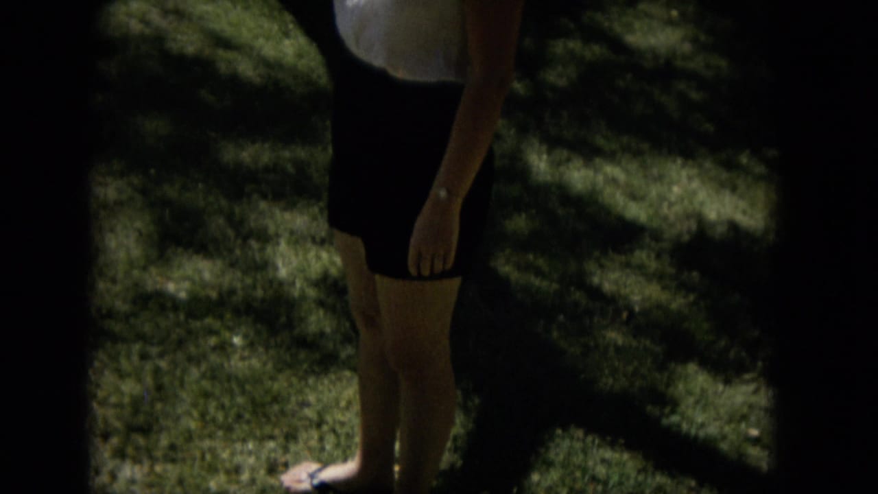 A dark contact-sheet band crosses the frame while the woman’s blouse, skirt, legs, and footwear remain visible against the shaded lawn in the 1965 home-movie footage.