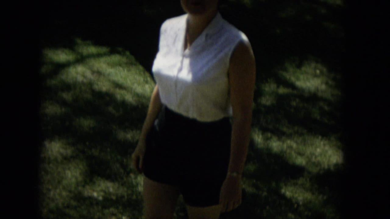 A woman in 1965 Hawaii home-movie footage walks through sun-dappled greenery wearing a white sleeveless blouse and dark skirt, with her face partly cropped by the frame.