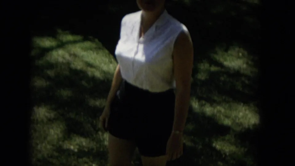 A woman in 1965 Hawaii home-movie footage walks through sun-dappled greenery wearing a white sleeveless blouse and dark skirt, with her face partly cropped by the frame.
