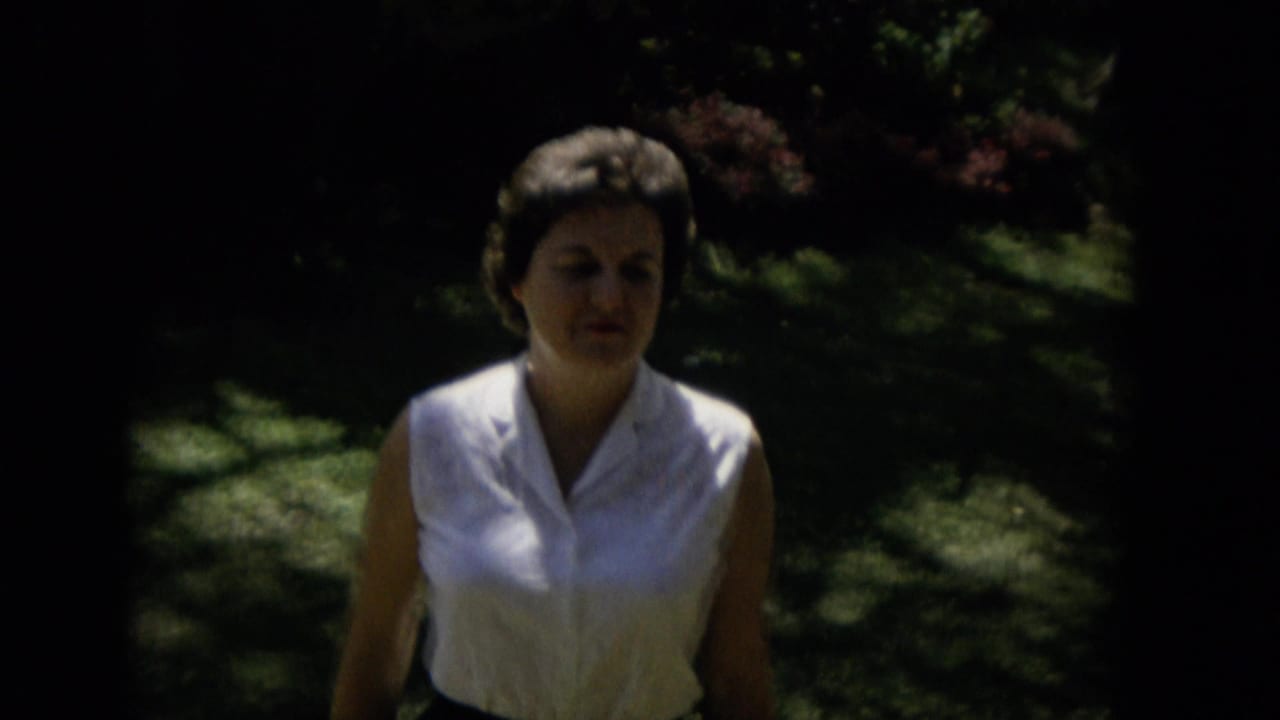 A woman in a white sleeveless blouse and dark skirt appears closer to the camera in shaded outdoor greenery, with strong contrast from the home-movie exposure.