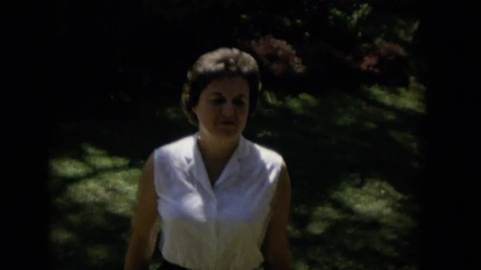 A woman in a white sleeveless blouse and dark skirt appears closer to the camera in shaded outdoor greenery, with strong contrast from the home-movie exposure.