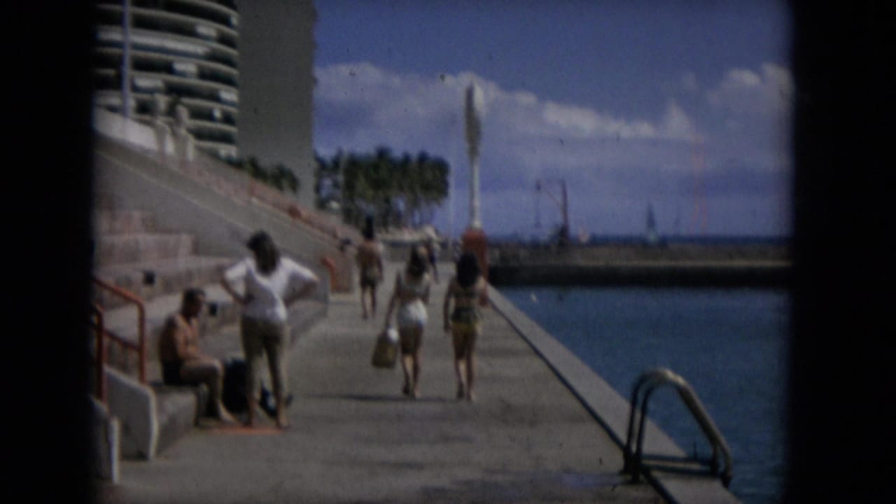 People stroll along a concrete waterfront promenade in Hawaii, with calm blue water, palm trees, a high-rise building, and a vertical fountain-like feature visible in the distance.
