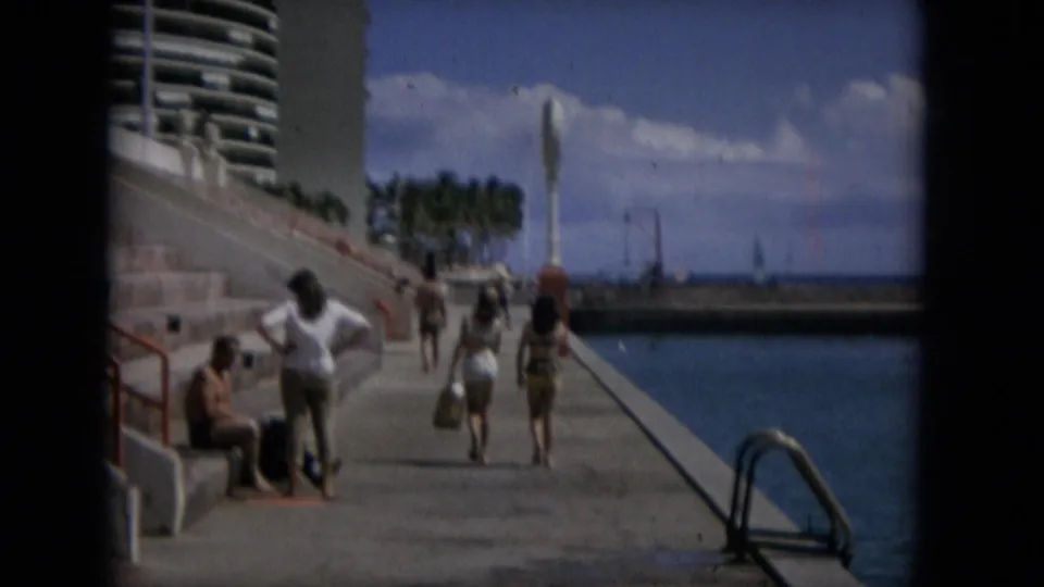 People stroll along a concrete waterfront promenade in Hawaii, with calm blue water, palm trees, a high-rise building, and a vertical fountain-like feature visible in the distance.