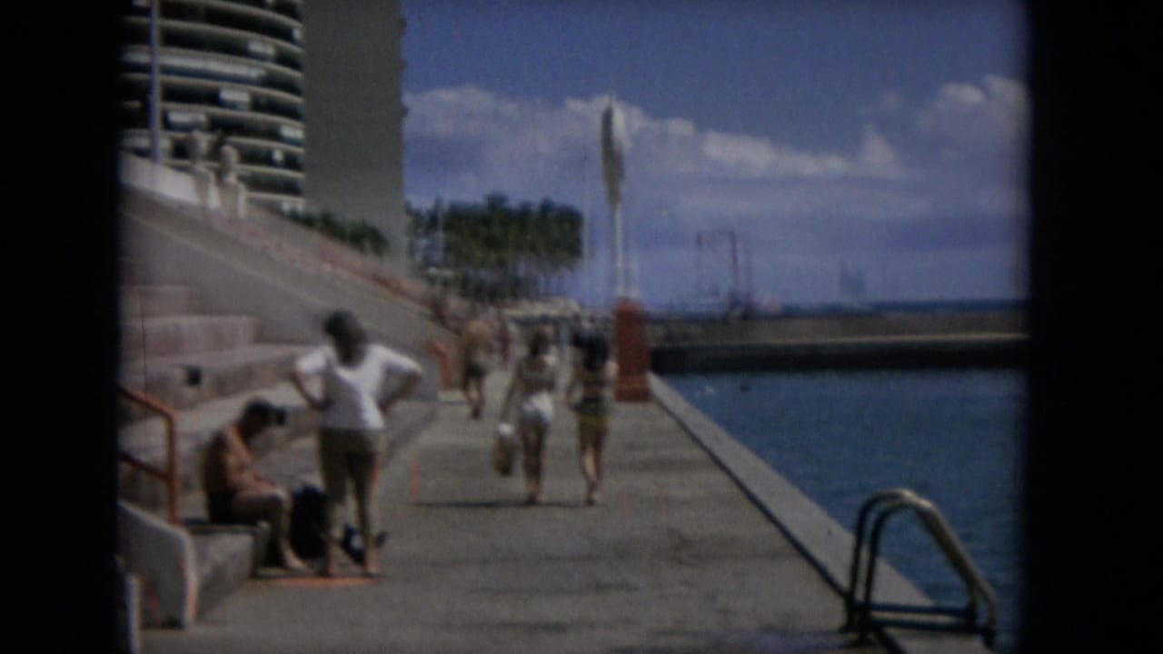 A later frame shows the same waterfront promenade with walkers and seated visitors beside blue water, palm trees, and a modern high-rise building under a bright sky.