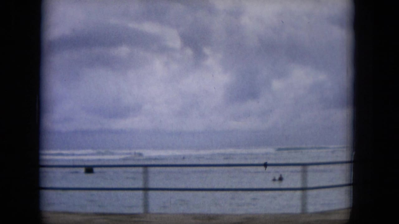 1965 Hawaii home-movie frame showing a cloudy ocean horizon beyond a seaside railing, with small people visible near the waterfront.