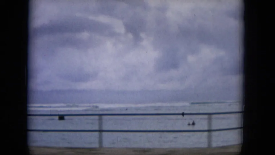 1965 Hawaii home-movie frame showing a cloudy ocean horizon beyond a seaside railing, with small people visible near the waterfront.