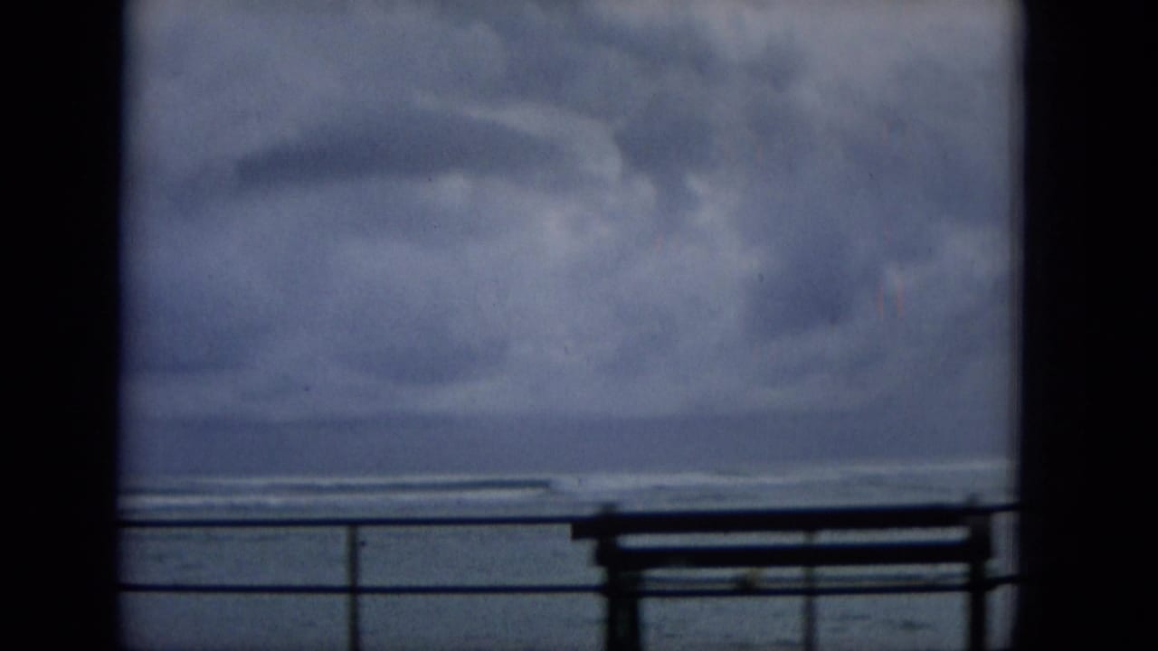 Soft 8mm frame from 1965 Hawaii showing a waterfront bench and railing beneath a broad cloudy sky over the sea.