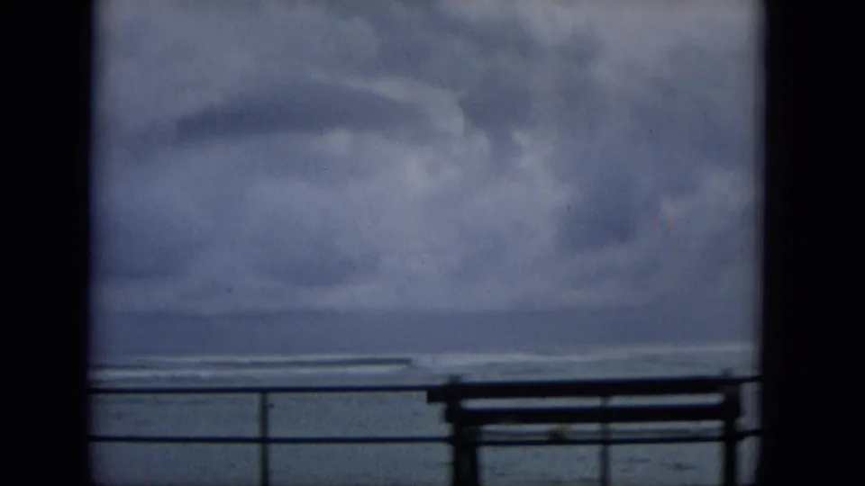 Soft 8mm frame from 1965 Hawaii showing a waterfront bench and railing beneath a broad cloudy sky over the sea.