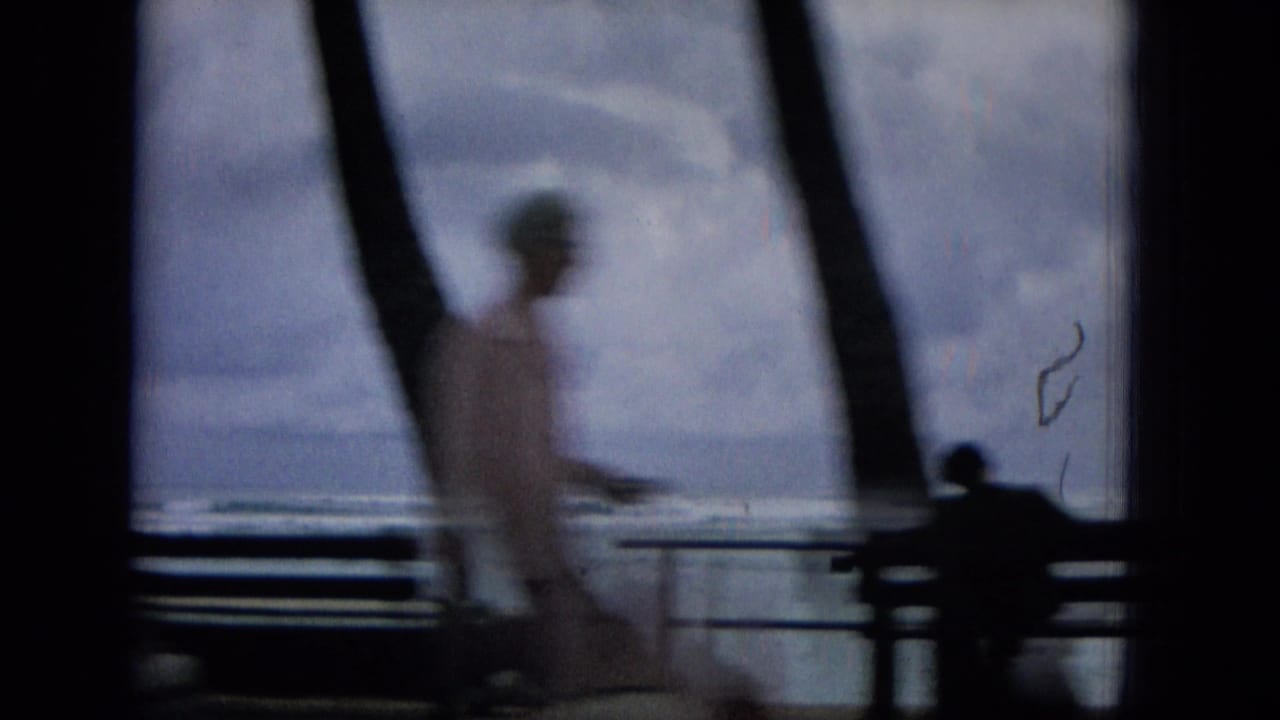 1965 Hawaii home-movie frame with a blurred person walking across a seaside promenade, benches and railings in front of a cloudy ocean view.