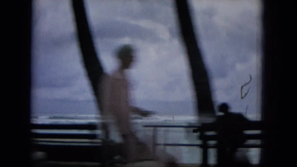1965 Hawaii home-movie frame with a blurred person walking across a seaside promenade, benches and railings in front of a cloudy ocean view.