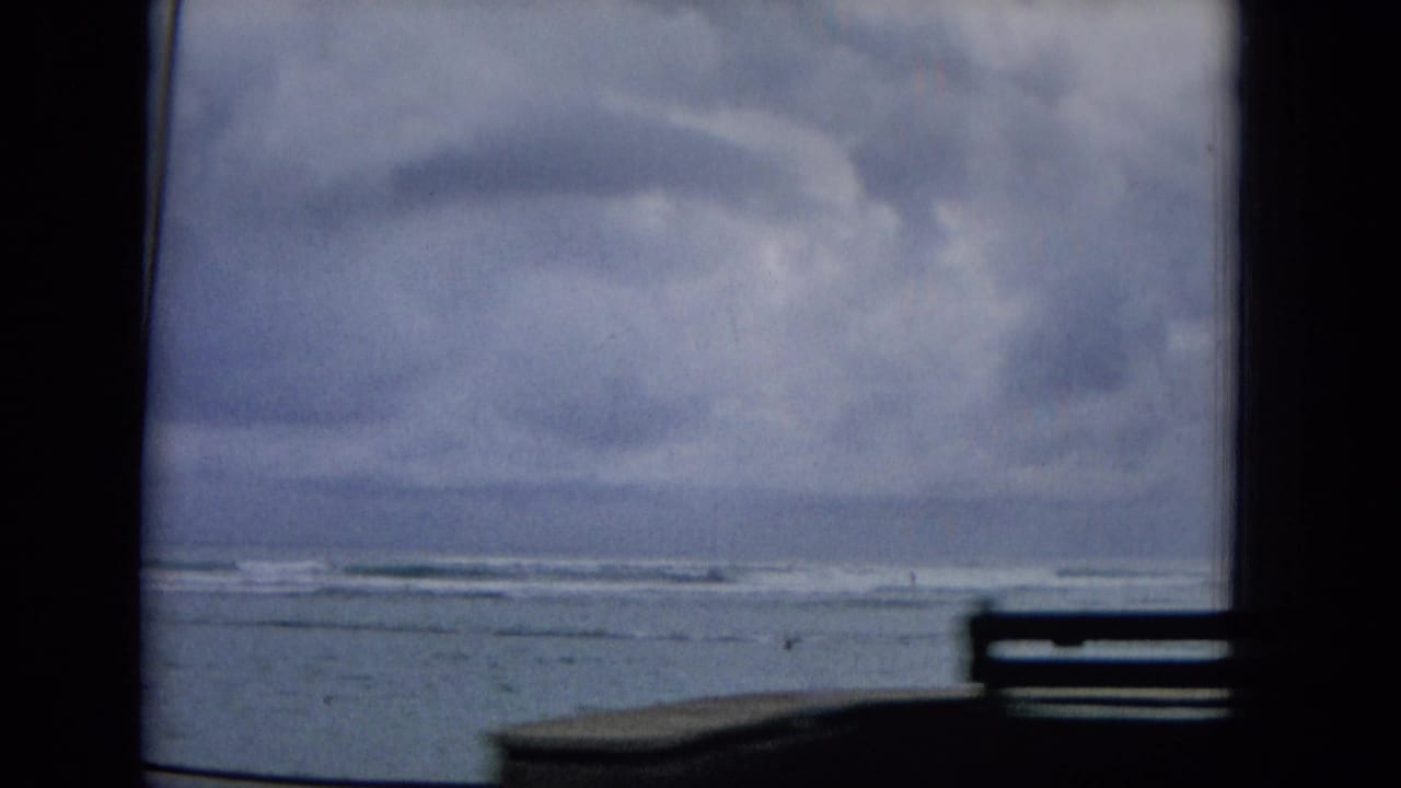 1965 Hawaii 8mm home-movie frame showing gray clouds, rough coastal water, and a distant shoreline through the dark interior framing of a moving vehicle.