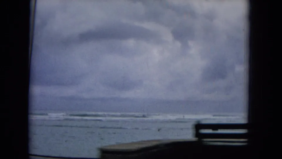 1965 Hawaii 8mm home-movie frame showing gray clouds, rough coastal water, and a distant shoreline through the dark interior framing of a moving vehicle.