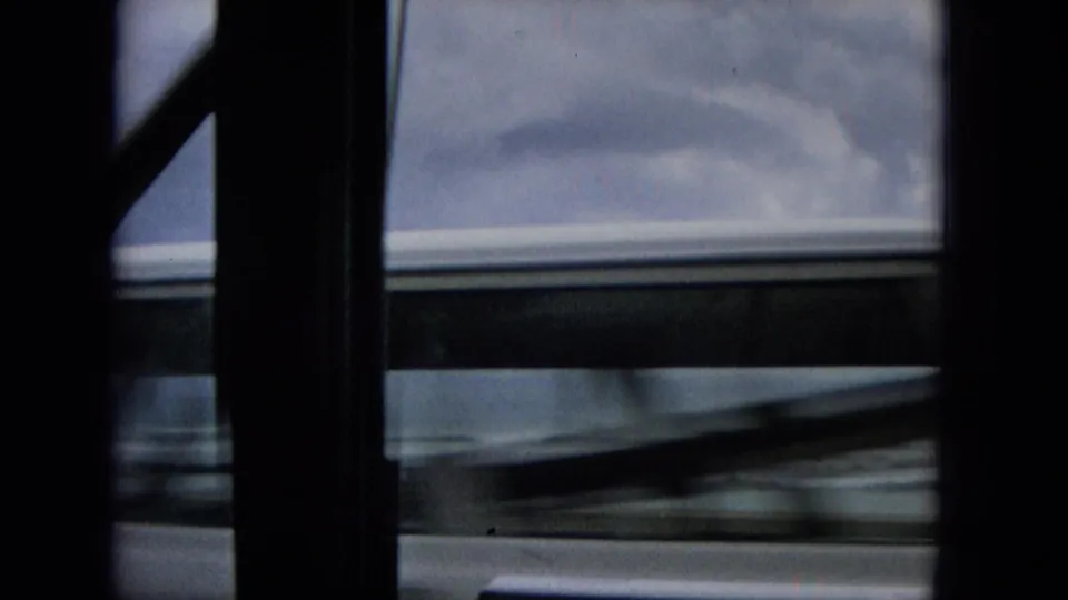 1965 Hawaii travel footage frame with dark window supports, a blurred passing vehicle or roofline, gray sky, and a partial coastal horizon seen from inside a moving vehicle.