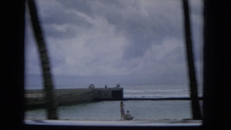 1965 Hawaii home-movie frame showing a beach, ocean surf, palm trunks, a low pier or seawall, and distant people along the shoreline from a moving vehicle.