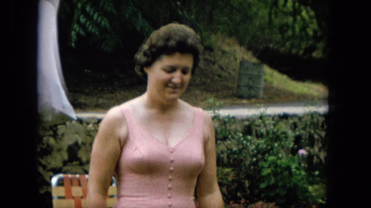 A woman in a pink button-front bathing suit stands outdoors in a Hawaii garden setting in 1965, with greenery, a stone wall, and a striped folding chair behind her.