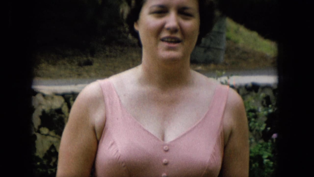 Closer 1965 home-movie view of the woman in a pink bathing suit looking toward the camera, with a stone wall and garden background visible behind her.